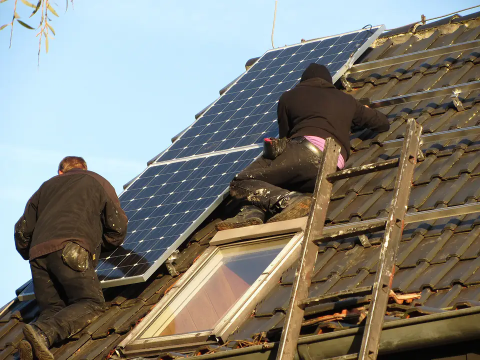 Deux artisans posent des panneaux photovoltaïques sur une toiture en tuiles, l’un sur une échelle de toit.